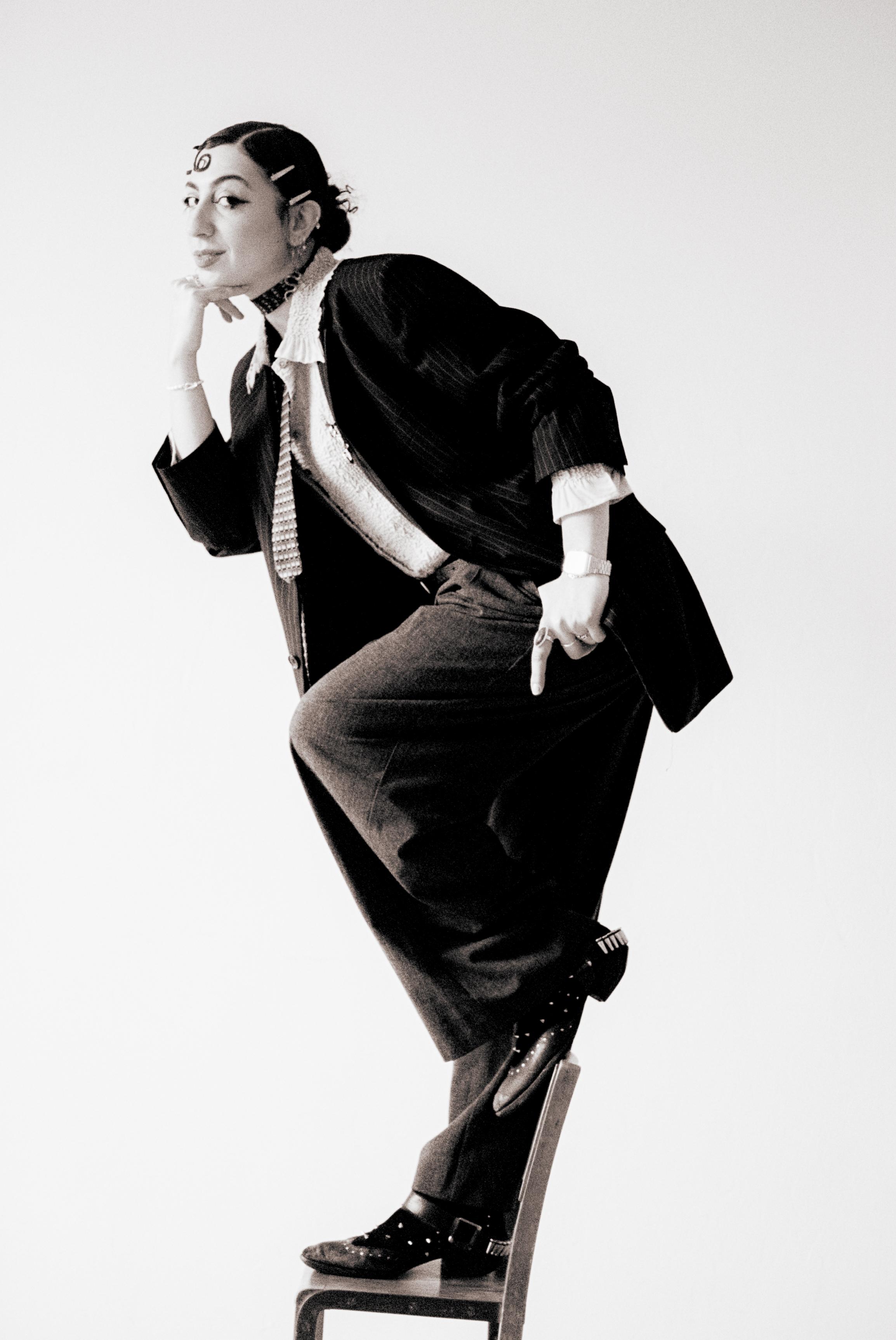 Rivkala seated in a tailored black suit against a pale studio background
